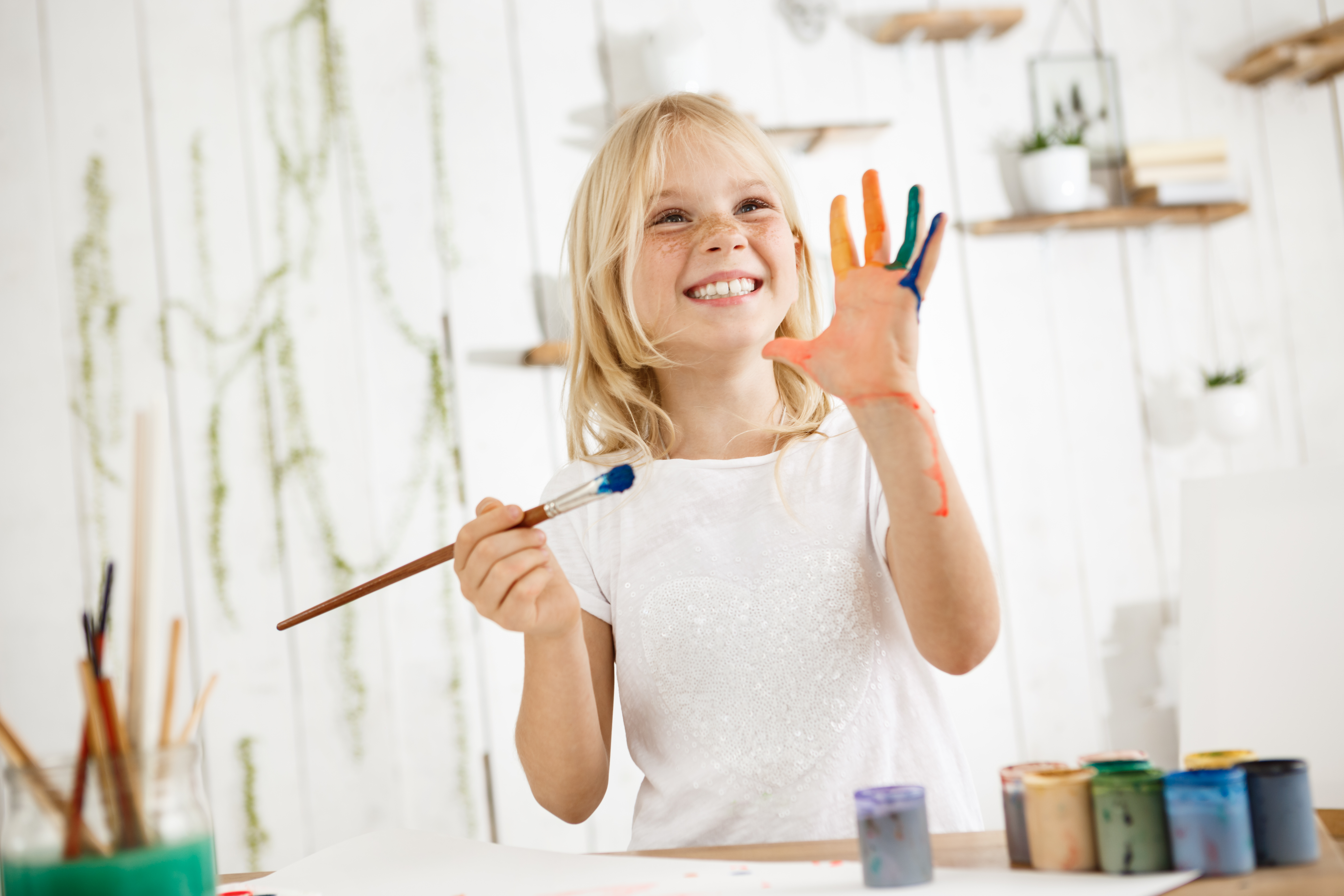 Happy and playful cute freckled blonde girl dressed in white, holding brush in one hand and showing another hand, which she messed up with paint. Children and happiness concept. Isolated shot, horizontal